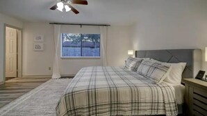 Une chambre à coucher avec un lit, un ventilateur de plafond, une fenêtre avec des stores et une table de chevet avec une lampe.