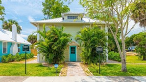 A two-story building with a teal door and windows, surrounded by lush greenery and palm trees.