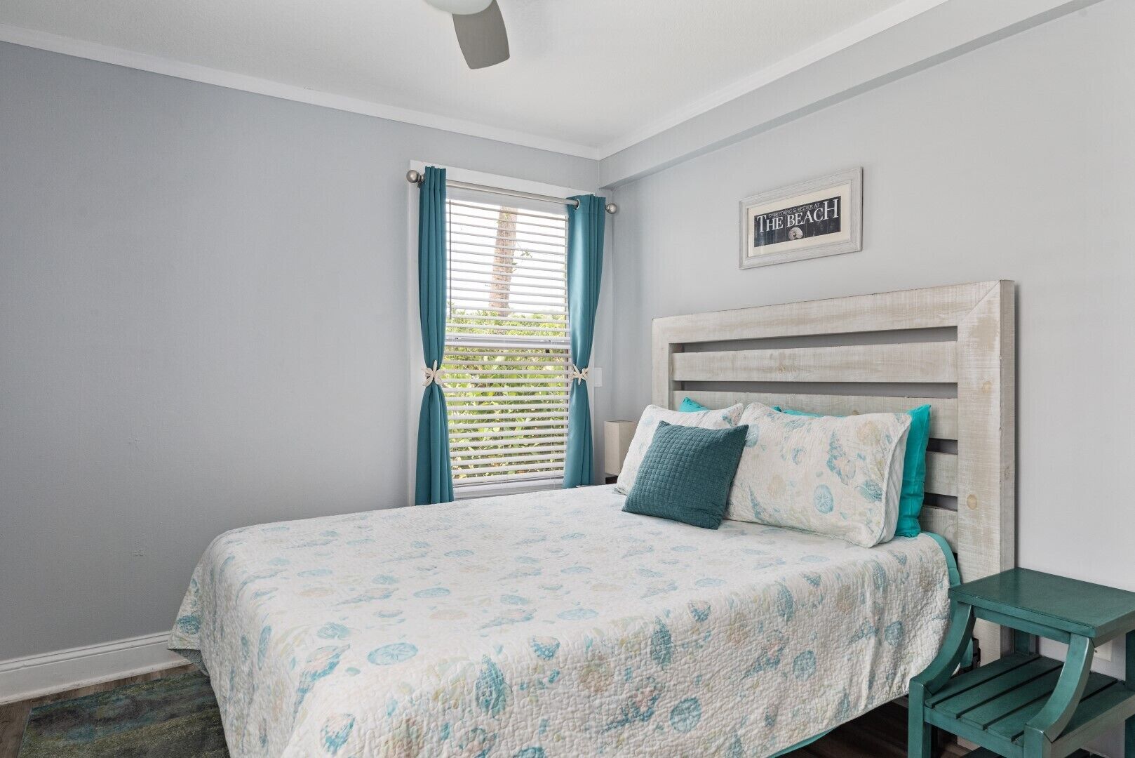A bedroom with a bed, a nightstand, a window with curtains, and a wall sign.