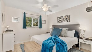 Un dormitorio con cama, mesita de noche, cómoda, ventilador de techo, ventana con cortinas y una obra de arte en la pared.