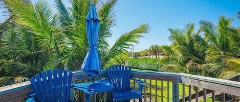 An outdoor deck with blue Adirondack chairs and a matching umbrella, surrounded by lush palm trees.