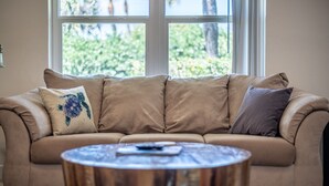 A beige sofa with multiple cushions, a round wooden coffee table, and a window with a view of greenery. - Tranquility Starfish Suite - Oceanfront Paradise! (Melbourne Beach)