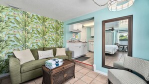 A living room with a green sofa, a coffee table, and a patterned wall. There is a kitchen visible through an open doorway. - Crane Creek Inn Dolphin Suite: Waterfront Pool! (Melbourne)