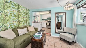 A cozy living room with a green sofa, a patterned armchair, and a coffee table with folded towels. A kitchen is visible in the background with white cabinets and a tiled backsplash. - Crane Creek Inn Dolphin Suite: Waterfront Pool! (Melbourne)