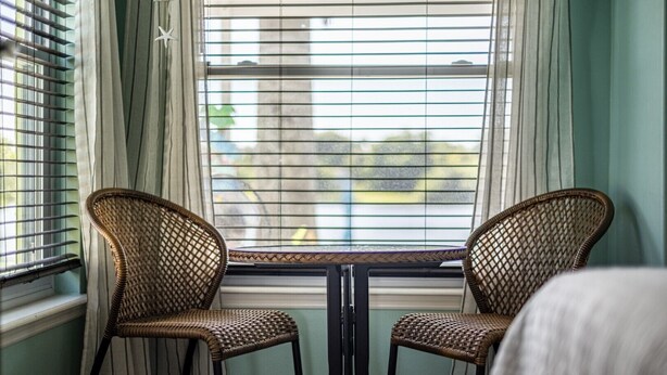 A room with two wicker chairs and a small table, a window with blinds, and a view of the outside. - Crane Creek Inn Dolphin Suite: Waterfront Pool! (Melbourne)