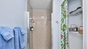 A bathroom with a glass shower door, a tiled shower floor, a towel rack with blue towels, and a shelf with toiletries and a basket. - Crane Creek Inn Manatee Suite: Waterfront Pool! (Melbourne)