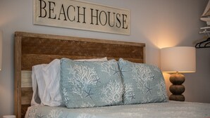 A bedroom with a wooden headboard, a bed with a blue and white patterned bedspread, a pillow with a coral reef design, a bedside table with a lamp, and a sign above the bed that reads "BEACH HOUSE". - Crane Creek Inn Manatee Suite: Waterfront Pool! (Melbourne)