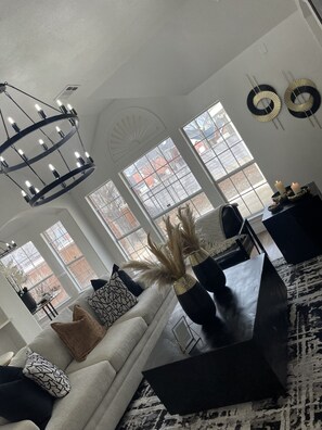 A modern living room with a chandelier, large windows, a sofa, and decorative items.