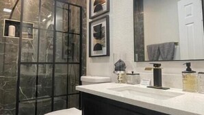 A modern bathroom with a glass shower enclosure, a white toilet, a black vanity with gold handles, and a large mirror above the sink.
