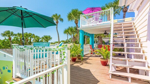 Una terraza de madera con barandilla blanca, un parasol verde y una estructura turquesa con un parasol púrpura.