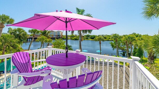 A deck with a purple table and chairs, a pink umbrella, and a view of a lake and palm trees.