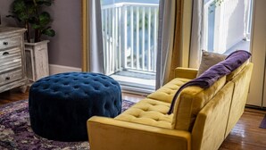 A room with a yellow sofa, a blue ottoman, and a view of a balcony with a railing.