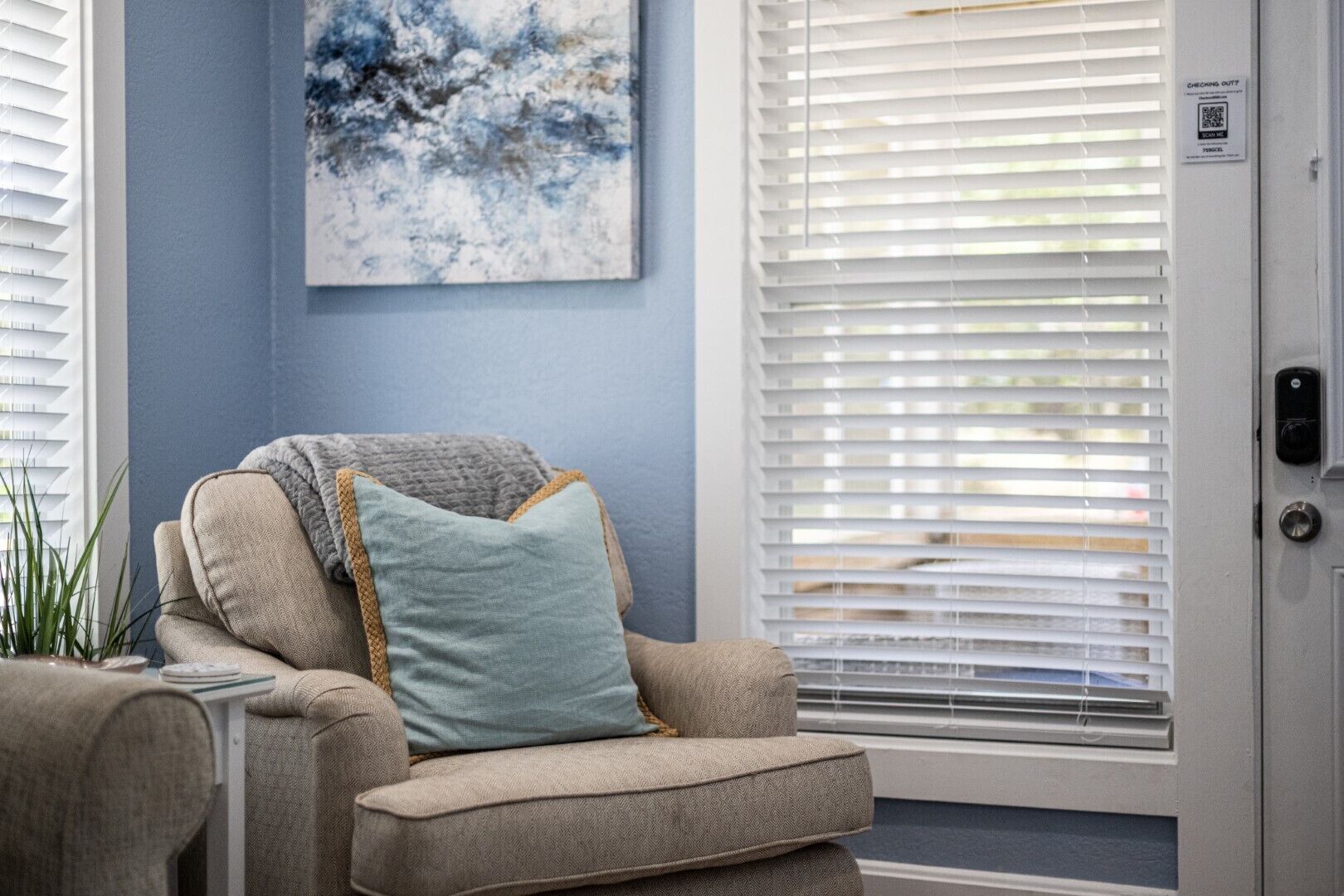Una habitación con un sillón color beige, un cuadro abstracto azul y persianas blancas.