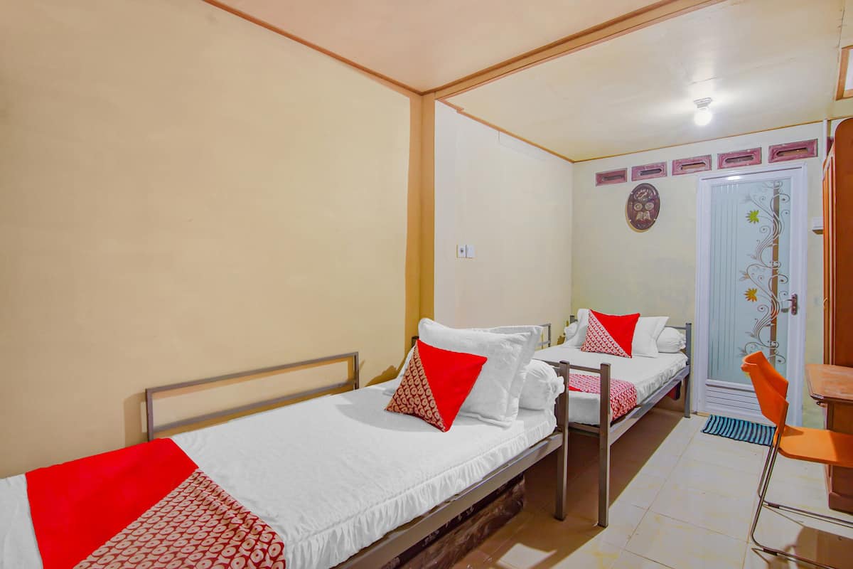 A small, well-lit room with two single beds, each with a red and white patterned bedspread. There is a wooden chair and a small table in the corner.