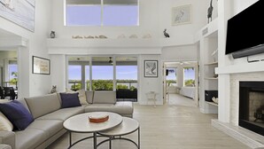 A spacious living room with a large sectional sofa, a round coffee table, and a fireplace. There are multiple windows offering a view of the outdoors.