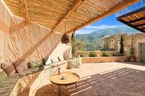 A cozy outdoor seating area with a wooden table, cushions, and a view of a historic stone building. - Villa Mirage - Private Plunge Pool & Views (Kournás)