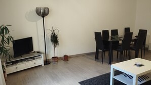 A modern living room with a flat-screen TV, a dining table with chairs, a floor lamp, and two potted plants.