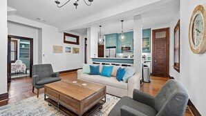 A spacious living room with a sofa, two armchairs, and a coffee table. A kitchen with teal cabinets and a dining area with a table and chairs are visible in the background. - Historic Loft in the Heart of Downtown (Galveston)