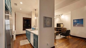 A modern kitchen with a central island, wooden flooring, and a dining area with a dark chair and a TV. - Historic Loft in the Heart of Downtown (Galveston)