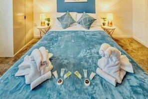 A hotel room with a bed, two bedside tables with lamps, a blue textured rug, and towels arranged on the rug. - The Sale Luxe | Captivating 2BR w/ Secured Parking (Manchester)