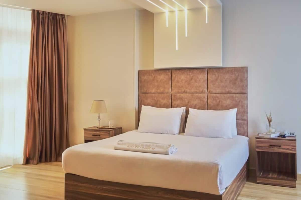 Una habitación de hotel moderna con una cama grande, suelo de madera, cabecera con iluminación integrada, mesita de noche con lámpara y una ventana con cortinas.