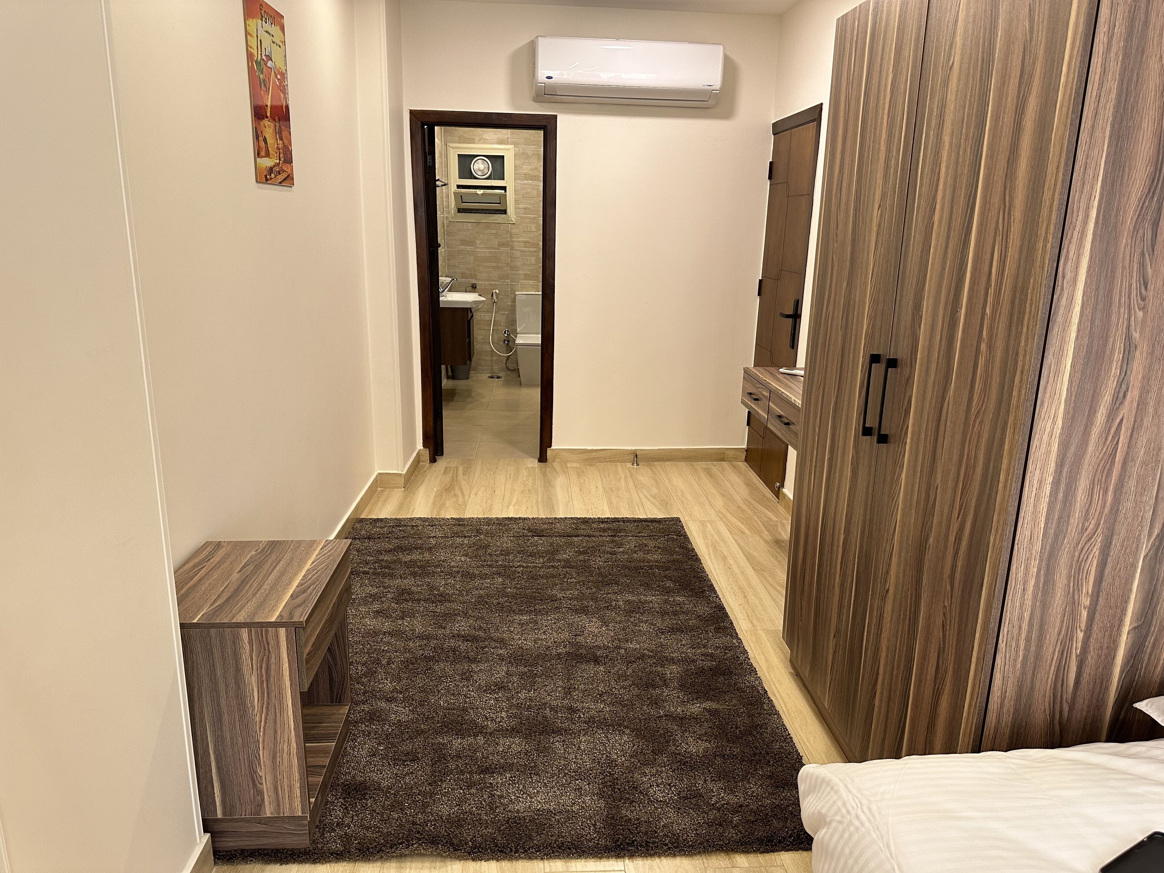 Un couloir étroit avec une armoire en bois, un tapis sombre et un lit recouvert de draps blancs.