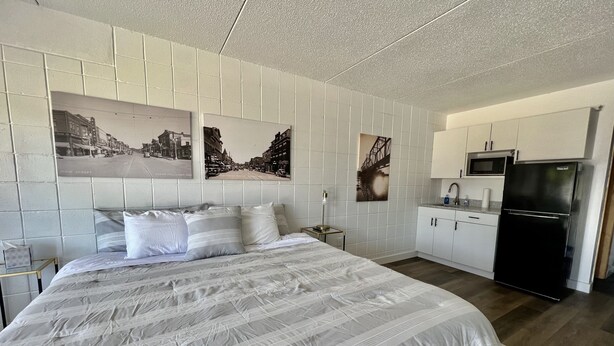 A bedroom with a large bed, a nightstand, a wall-mounted lamp, and a kitchen area with a refrigerator and cabinets.