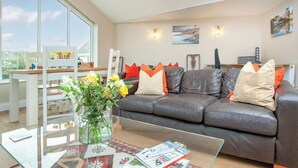 A modern living room with a glass coffee table, a leather sofa with orange and white cushions, and a large window offering a view of the outdoors.