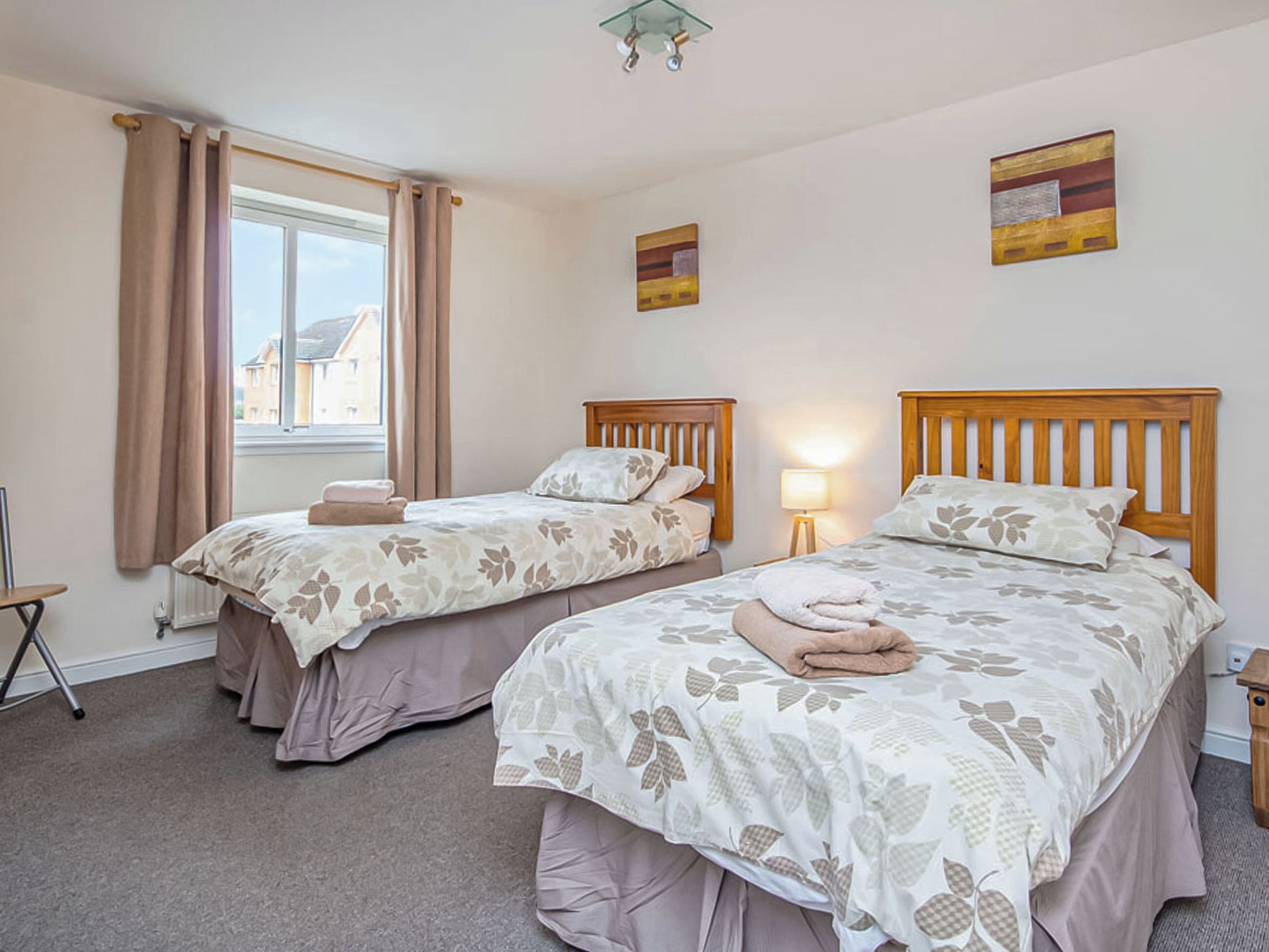 A hotel room with two beds, a window with curtains, a lamp, and artwork on the walls.