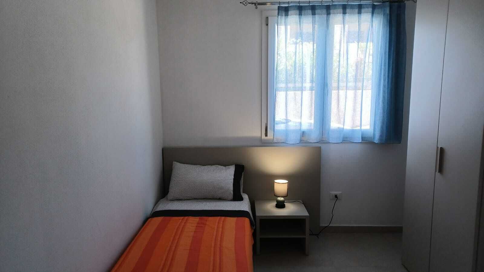 Una habitación pequeña con cama individual, mesita de noche, lámpara y ventana con cortinas de gasa.