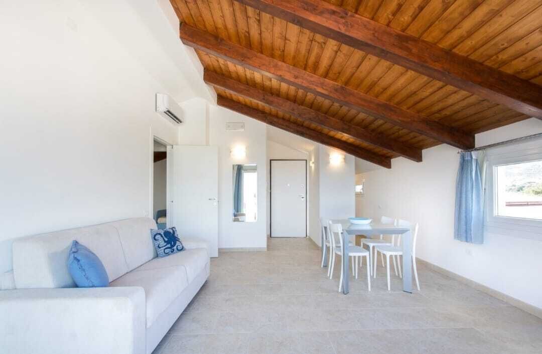 Un'ampia zona giorno con soffitto in legno a vista, un divano bianco con cuscini blu, un tavolo da pranzo con sedie bianche e una porta che conduce in un'altra stanza.