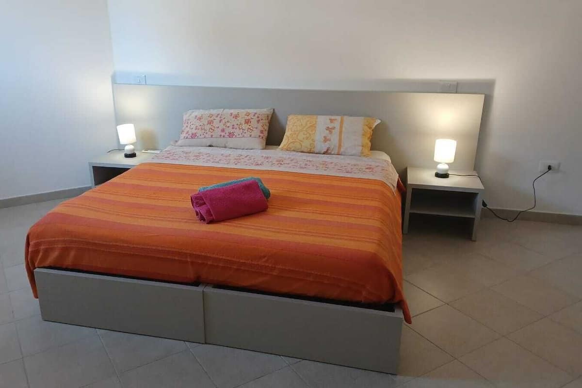 A neatly made bed with a striped orange bedspread and two pillows, flanked by bedside tables with lamps, in a simple room with plain walls and tiled flooring.
