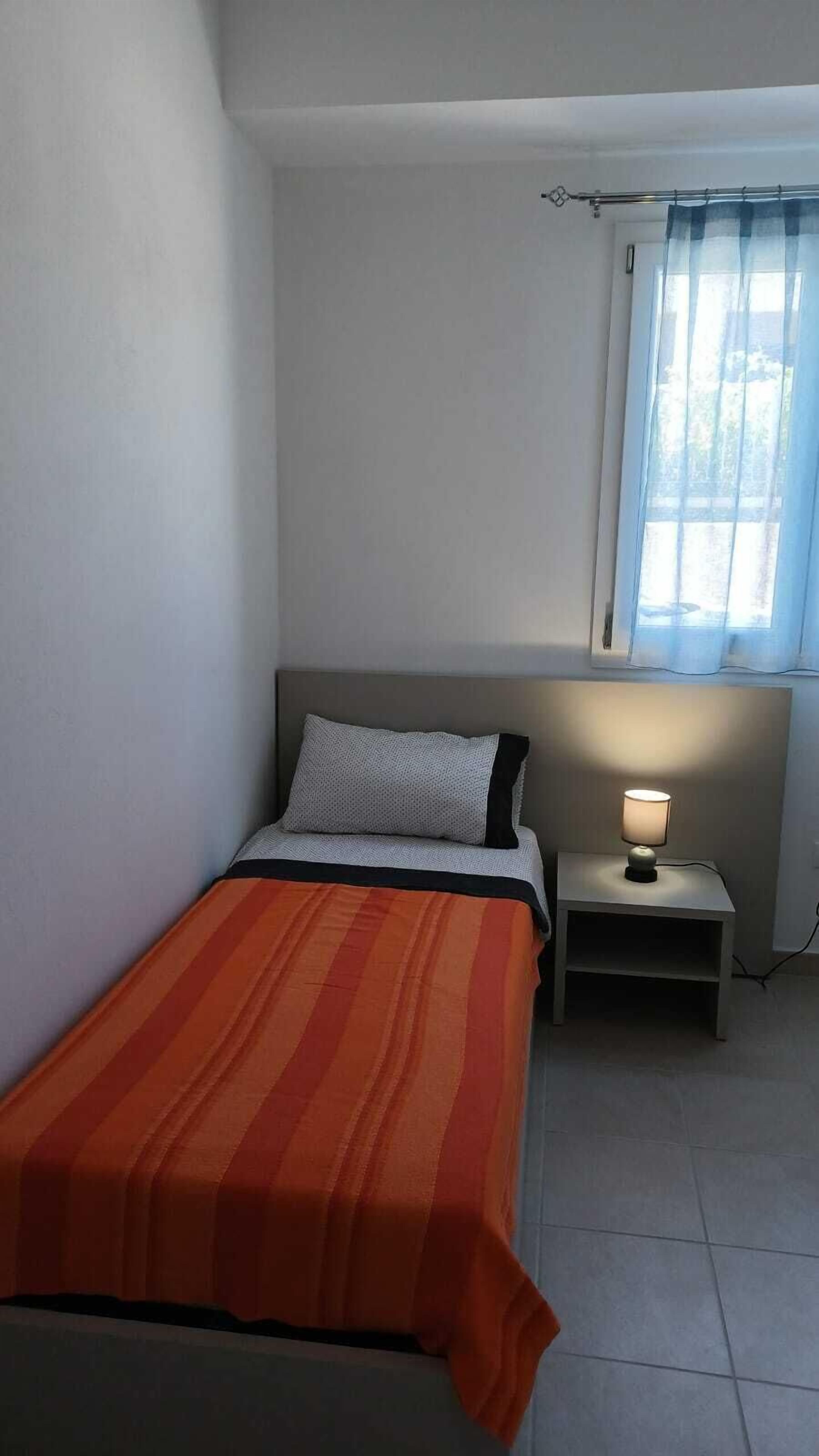 A small, simple bedroom with a single bed, a nightstand, a lamp, and a window with curtains.
