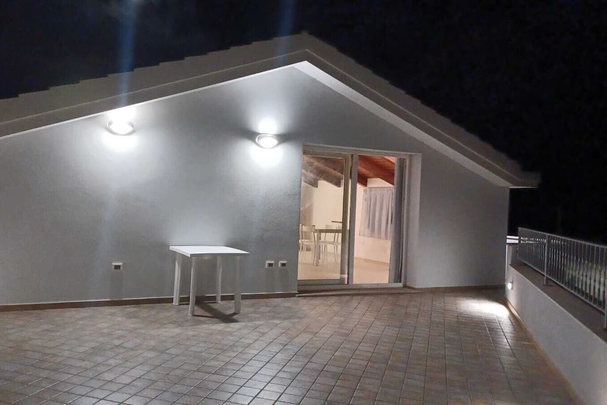 A covered outdoor area with a table and chairs, a tiled floor, and a white building with a sloped roof and two lights.