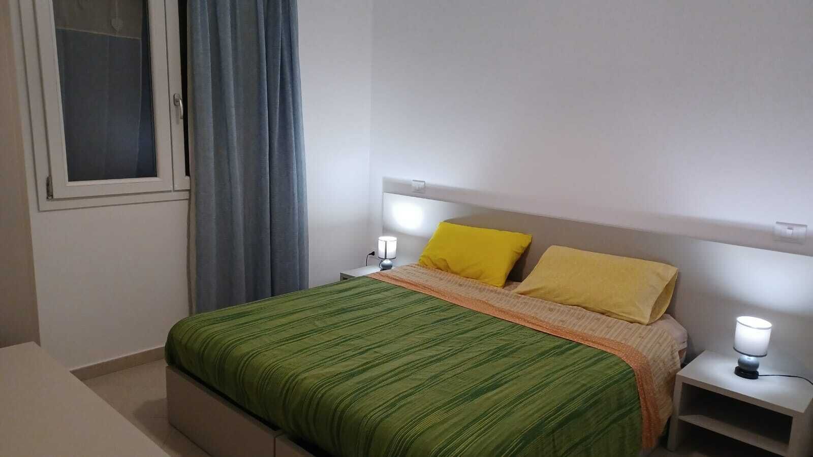 Un dormitorio con una cama, dos lámparas de noche, una ventana con cortinas y una mesita de noche.
