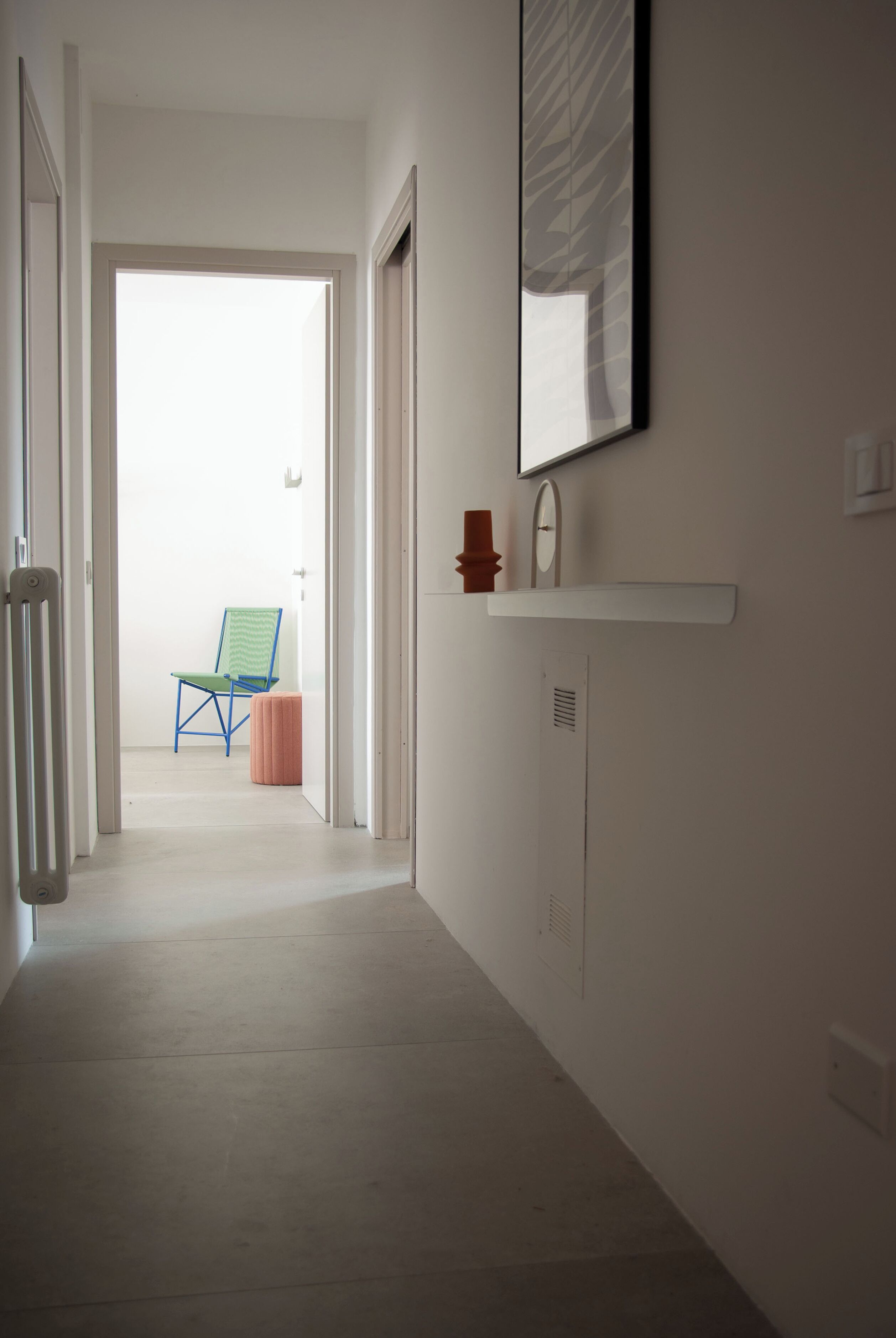 A hallway leading to a room with a chair and ottoman, a mirror, and a shelf with a small item.