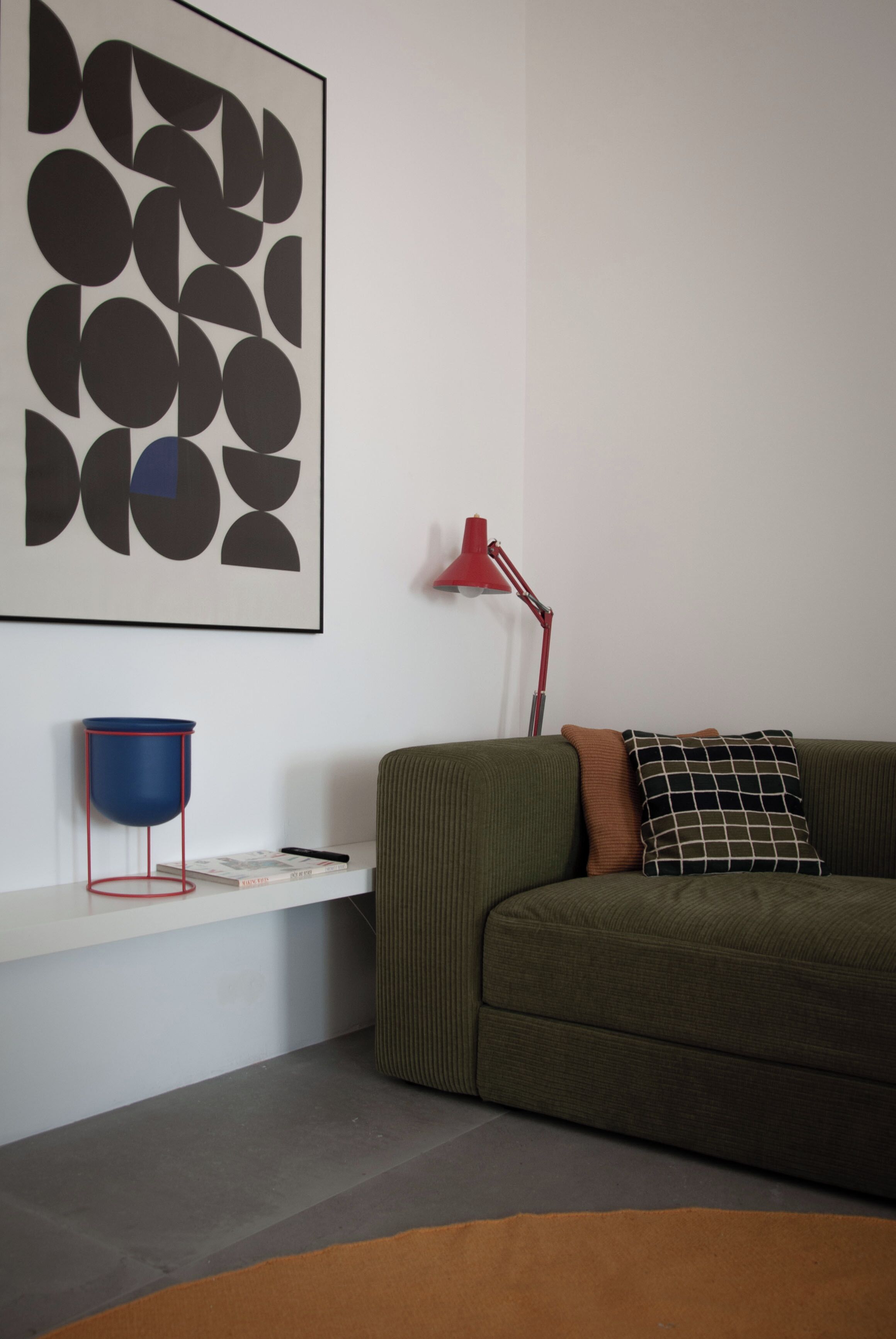 Un salon moderne avec un canapé vert, un lampadaire rouge, une œuvre d’art abstraite encadrée et un lampadaire bleu posé sur une étagère blanche.