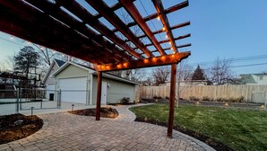 Un patio couvert avec un chemin en briques, une pergola en bois et des guirlandes lumineuses.