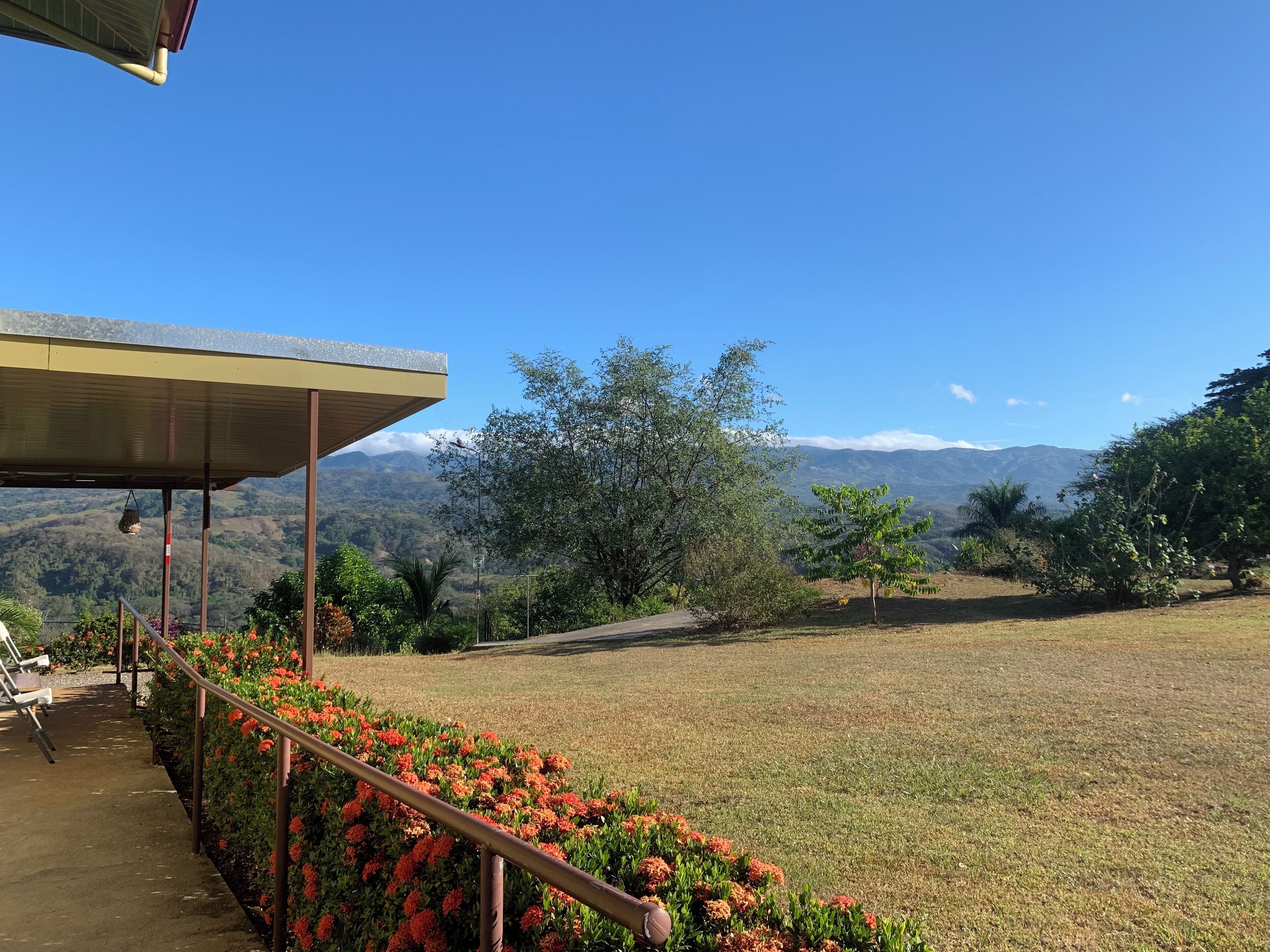 A covered outdoor area with a concrete pathway, benches, and a railing overlooking a grassy field with trees and distant mountains.