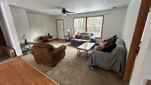 A spacious living room with a brown armchair, a grey sofa, and a small table. There is a window with a view of trees, and a ceiling fan. - NEW LISTING - FARMHOUSE --VERY CLOSE TO SNOWMOBILE TRAILS - VERY PRIVATE (Brimley)