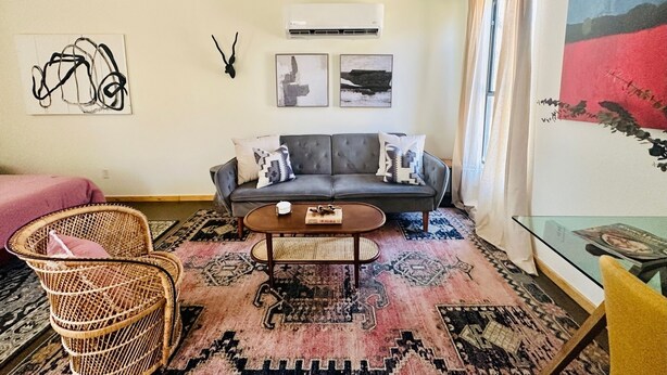 A living room with a grey sofa, two patterned cushions, a wicker chair, a coffee table, and a glass table with magazines. - Hill Country Bungalow at Mosaic (San Marcos)