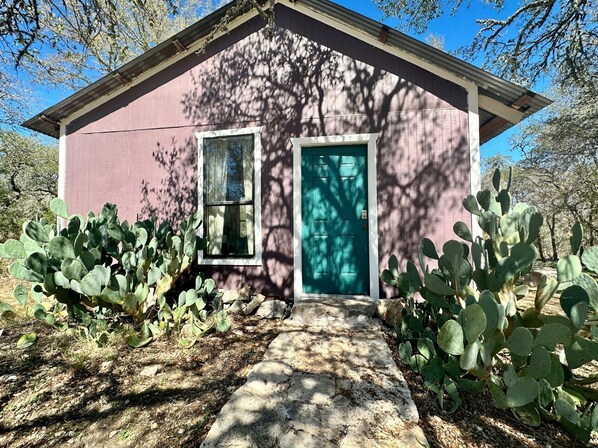 Una casita con puerta y ventanas color turquesa, rodeada de cactus.