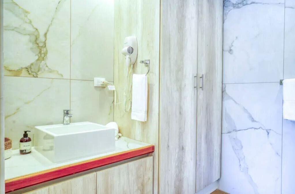 Um banheiro com parede revestida em mármore, pia branca com bancada vermelha e secador de cabelo fixado na parede.