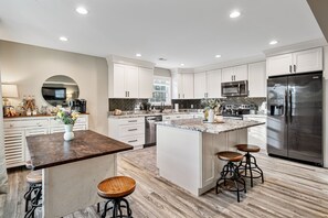 A modern kitchen with white cabinets, a large island with stools, stainless steel appliances, and a herringbone backsplash.