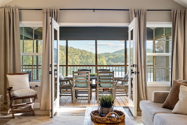 A room with a large sliding glass door leading to a balcony with a scenic view of trees and hills. Inside, there is a wooden table with chairs, a cushioned chair, and a sofa.