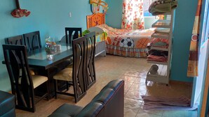 A room with a dining table, chairs, a bed with a colorful bedspread, and a shelf with folded clothes.