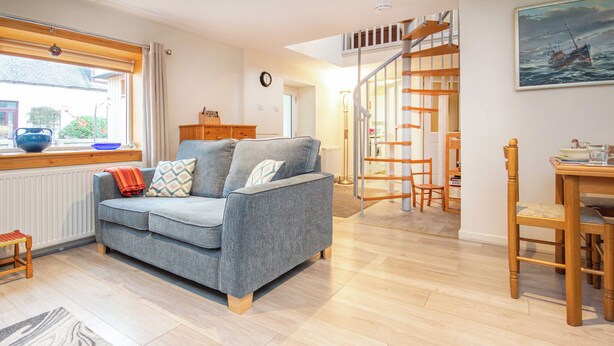 Een woonkamer met een grijze bank, houten vloer en een eethoek met een tafel en stoelen.
