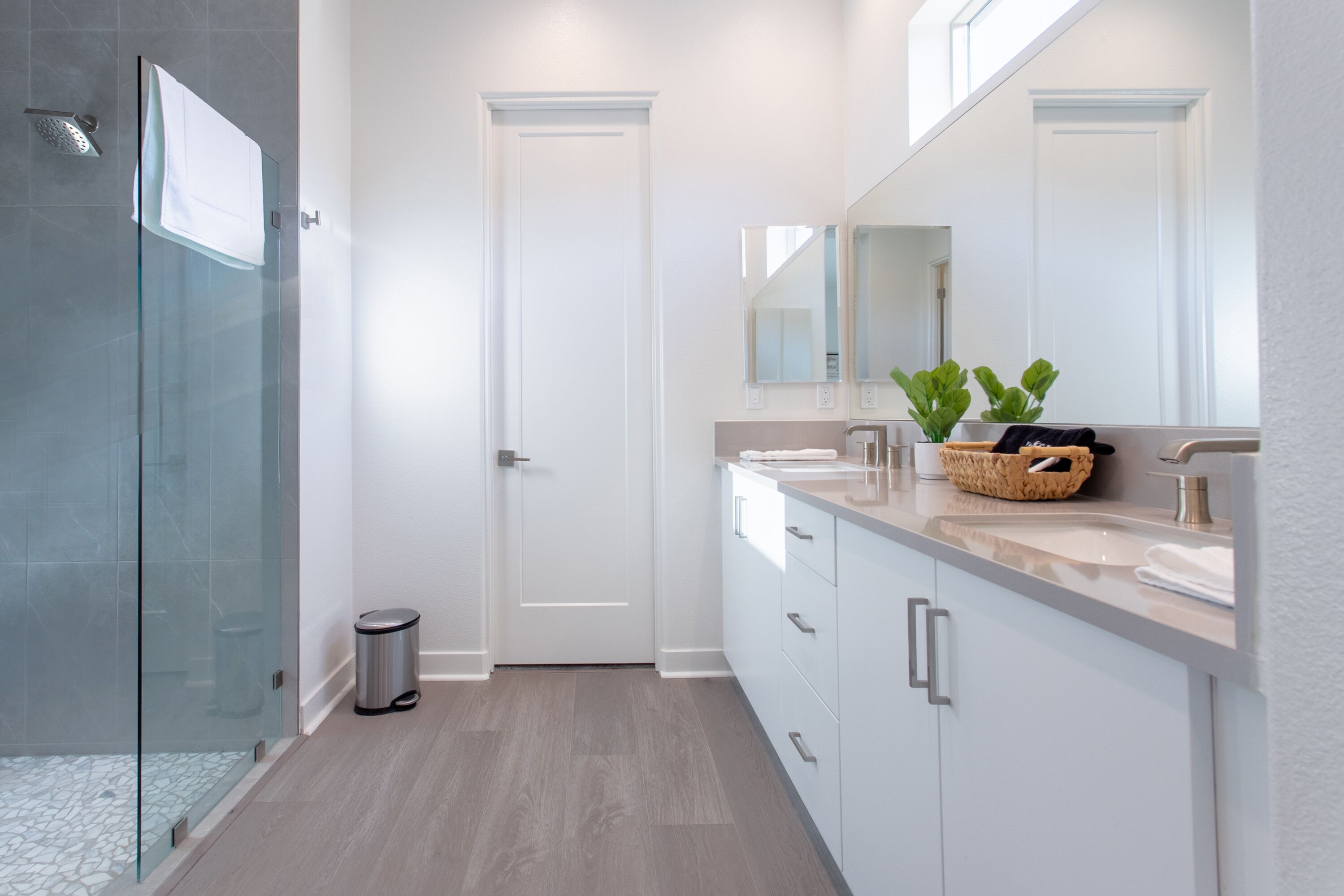 A modern bathroom with a glass shower enclosure, a white door, a sink with a mirror, and a basket of toiletries.