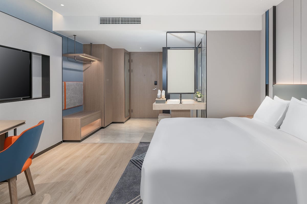 A modern hotel room with a large bed, a desk with a chair, a vanity with a mirror, and a TV mounted on the wall.