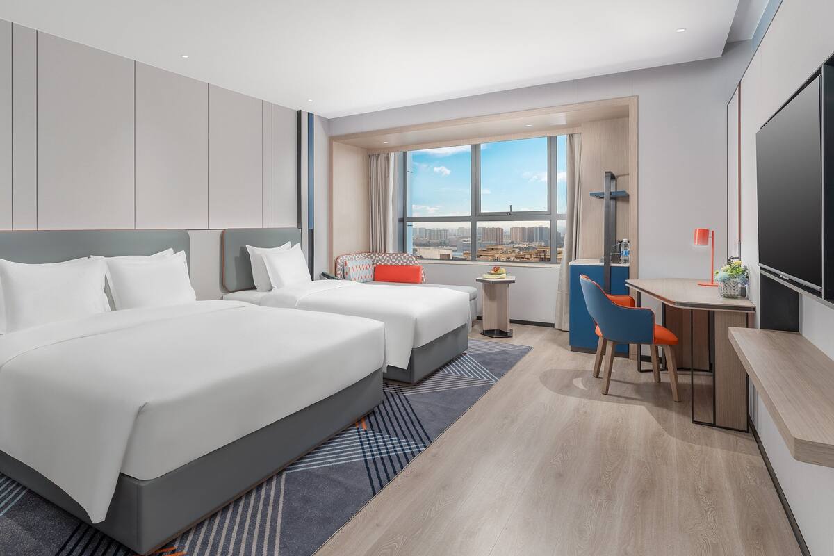 A modern hotel room with two beds, a desk, and a large window offering a city view.